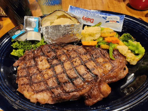 A grilled steak with a baked potato, sour cream, and mixed vegetables on a blue plate.